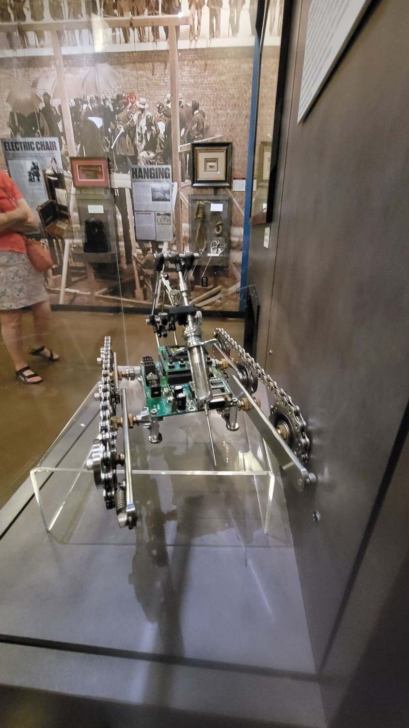 Figure 4 — Gallery context: the vitrine sits within the museum's survey of
execution methods, with HANGING and ELECTRIC CHAIR panels on the wall behind. The
placement frames the droid as the next s…